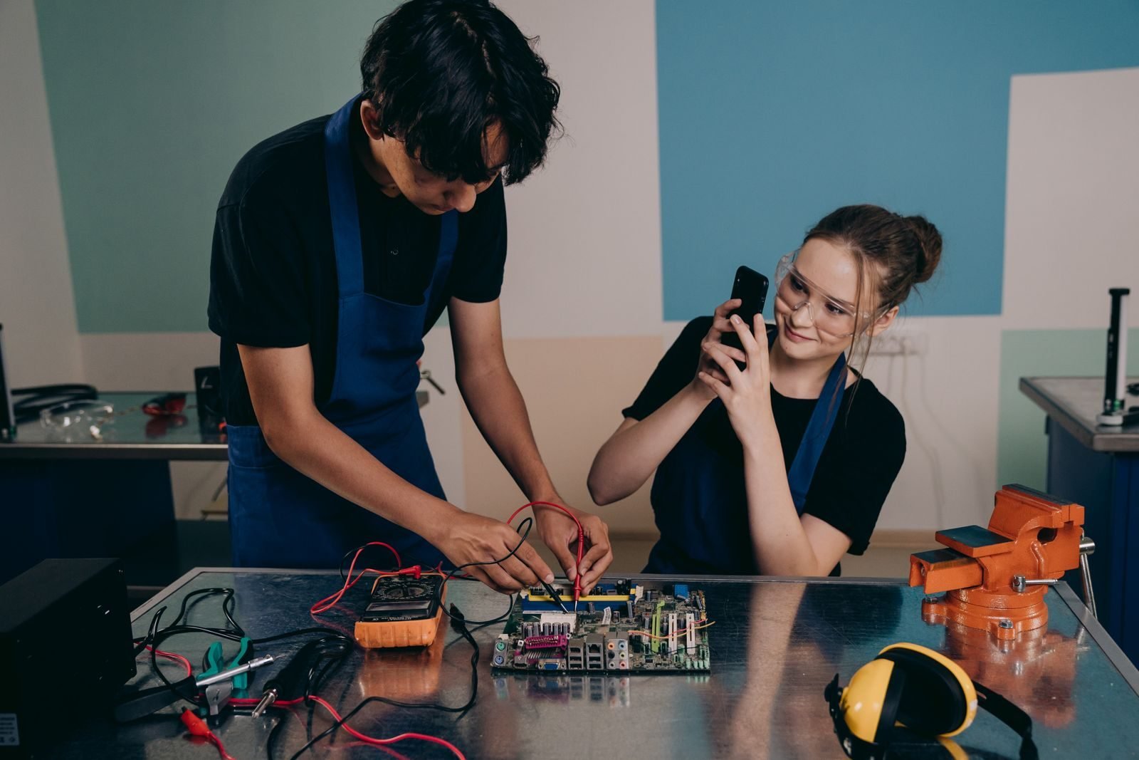 Two young adults testing a motherboard in a tech lab, capturing the process with a smartphone.